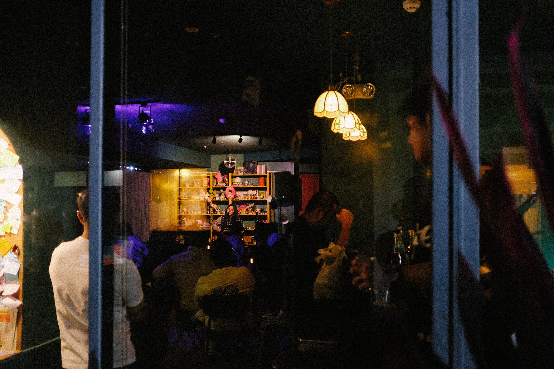 a dim night at Cattery, seen from the street — a full room watching a set, tungsten lamps glowing over the shelf of lucky cats