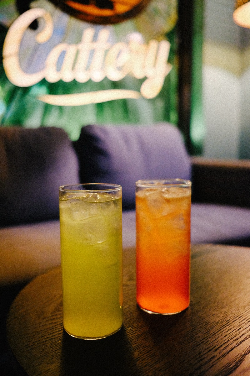 Two tall Cattery cocktails, one green and one orange over ice, on a wood table below the glowing Cattery sign