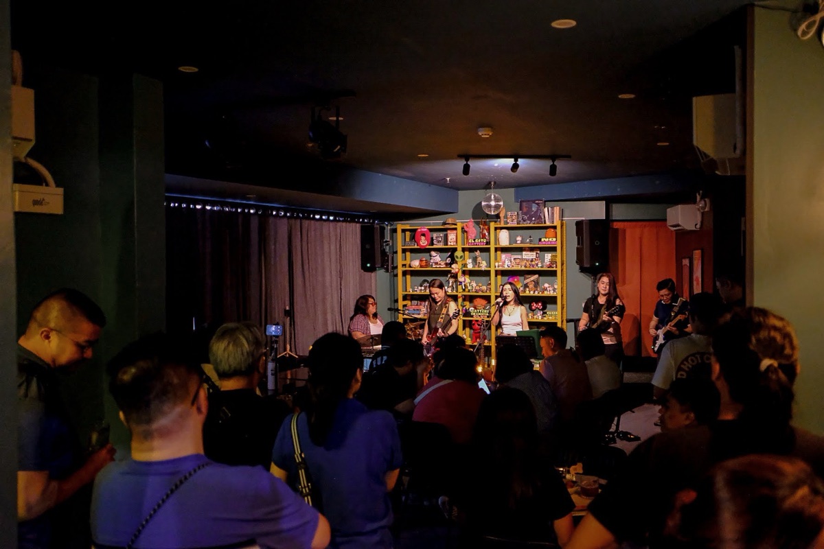A band mid-set at Cattery — singer and guitarists lit warm against the maneki-neko figurine shelf, a packed room watching up close