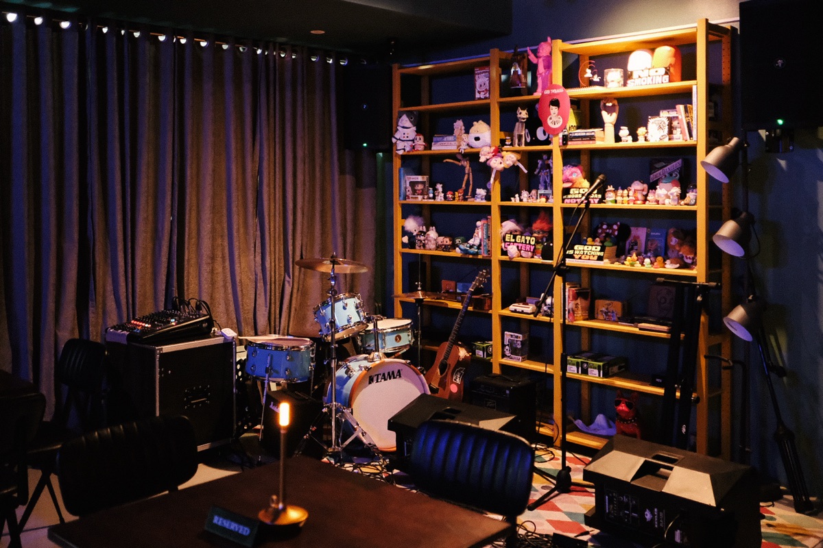Cattery's stage set and waiting — a blue drum kit, guitar, and amps beside the tall shelf of figurines and toys