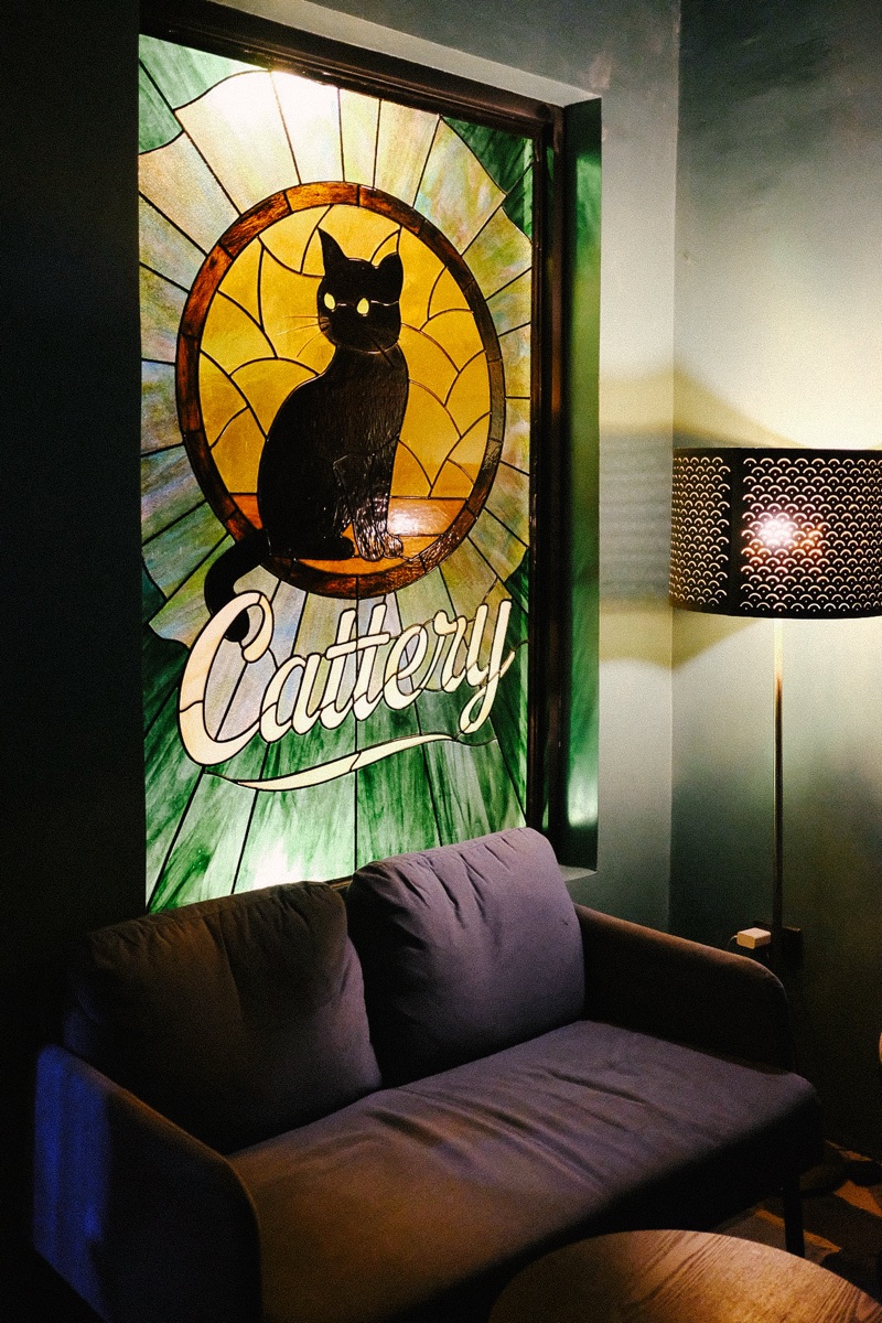 Cattery's stained-glass window — a black cat perched in a sunlit oval above the hand-lettered Cattery banner, beside a couch and a patterned floor lamp